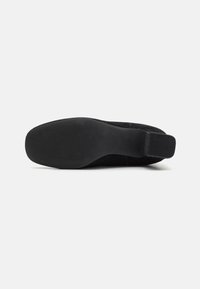 Zapato negro de tipo slip-on con parte superior de tejido texturizado, suela de goma plana y panel lateral elástico para un ajuste flexible. Superficie lisa y patrón sutil.