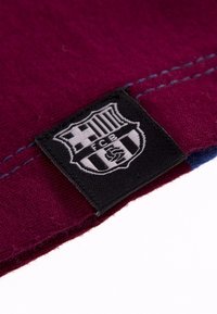 Bordeauxkleurig stoffen label met een zwart-wit FC Barcelona-logo. Zichtbare stiksels in een contrasterende lichtblauwe kleur.