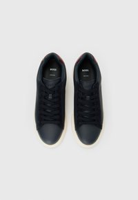 O pereche de adidași negri din piele, cu talpă albă și accente maronii la călcâi, marca "BOSS", afișați pe un fundal alb simplu.