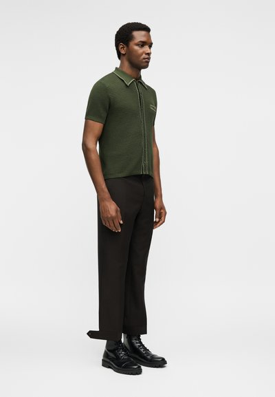 Άνδρας στέκεται φορώντας μια πλεκτή πόλο με κοντά μανίκια σε ελιά πράσινο, μαύρο κοντό παντελόνι και μαύρα μποτάκια με κορδόνια σε μονόχρωμο φόντο.