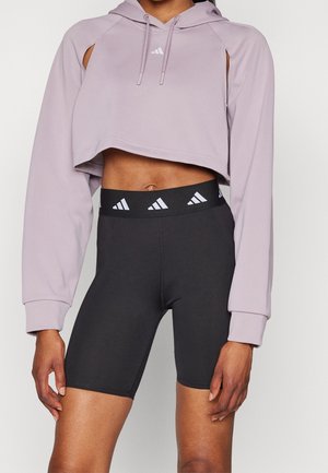 Środkowa część ciała osoby ubranej w lawendową, krótka bluzę z kapturem oraz czarne, wysokowalne krótkie spodenki rowerowe z białymi logo Adidas na pasku i na piersi.