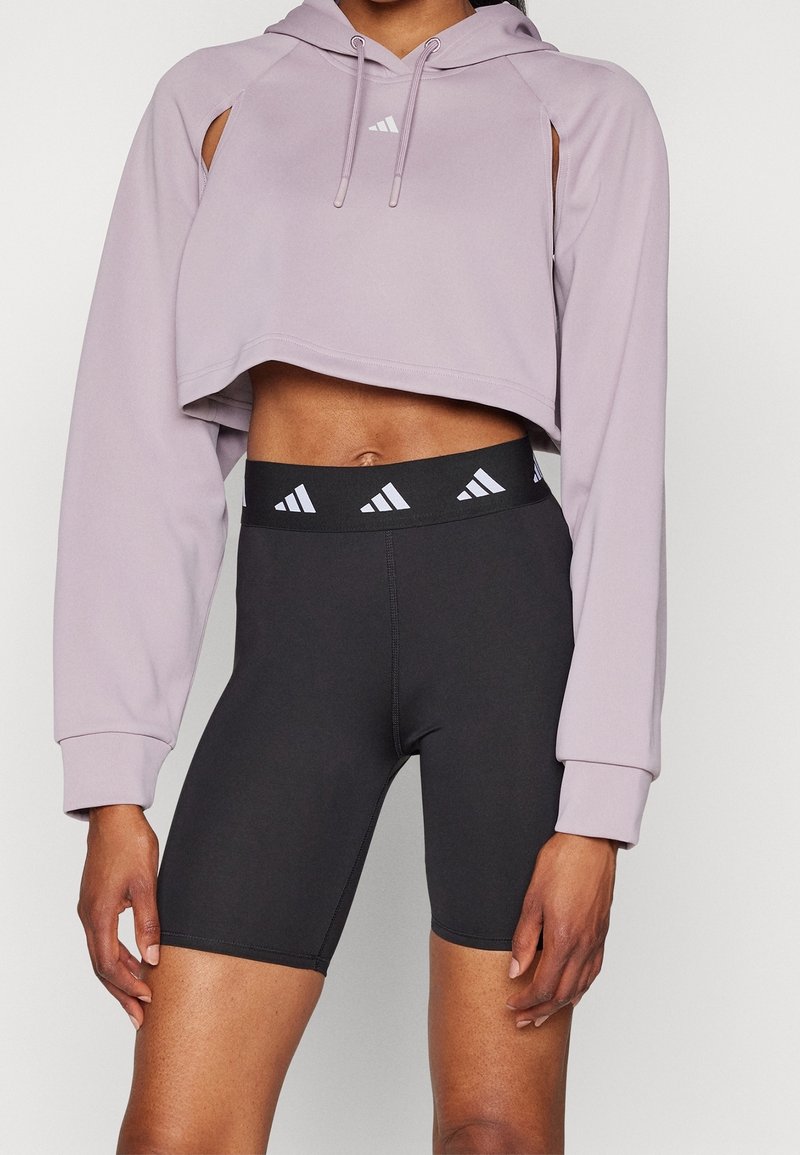 Torse d'une personne portant un sweat à capuche court lavande et un short de cycliste taille haute noir avec des logos Adidas blancs sur la ceinture et la poitrine.