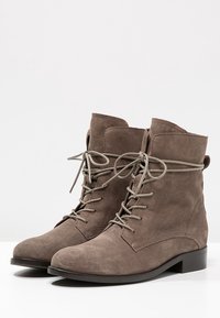 Braune Wildleder-Ankle-Boots mit runder Zehenpartie, Schnürungsfront und flacher Sohle. Das Design zeichnet sich durch eine weiche Textur und minimale Details aus.