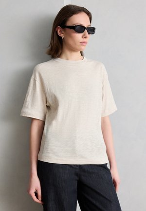 Mladý člověk nosící černé obdélníkové sluneční brýle, krémovou krátkou košili a tmavé kalhoty, stojící u světle šedé stěny.