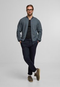 Strukturierte dunkelblaue Bomberjacke, schwarzes Shirt, marineblaue Hose und braune Schuhe. Das Outfit zeichnet sich durch eine lockere Passform und hochgerollte Hosenbeine aus.
