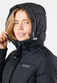 Schwarze Daunenjacke mit hohem Kragen und verstellbarem Kapuzen. Sie hat eine glänzende Verarbeitung, sichtbare Nähte und ein Logo auf der Brust.