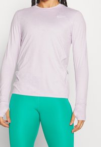 Jasnofioletowa sportowa bluzka z długim rękawem, okrągłym dekoltem i otworami na kciuki, z małym białym logo Nike na piersi.