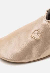 Metallisk gull slip-on sko med en jevn tekstur og rund tå; har en sydd hjerteaksent på siden. Mykt indre fôr.