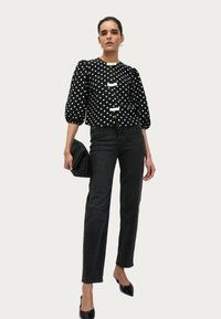 Blouse noire à pois blancs avec des manches bouffantes, ornée de trois boutons blancs, associée à un jean en denim noir taille haute et des escarpins noirs à bout pointu.