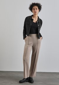 Zwarte getextureerde jas met borstzakken, gedragen over een zwart topje. Broek in lichtbeige met verticale naden, gecombineerd met zwarte enkellaarsjes.