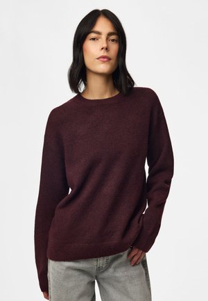 Femme aux cheveux noirs mi-longs, portant un pull violet foncé et un jean gris clair, debout devant un fond blanc uni.