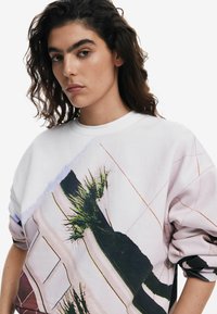 Sudadera blanca con un gran estampado abstracto de formas y acentos verdes. El diseño incluye líneas diagonales y una textura suave.