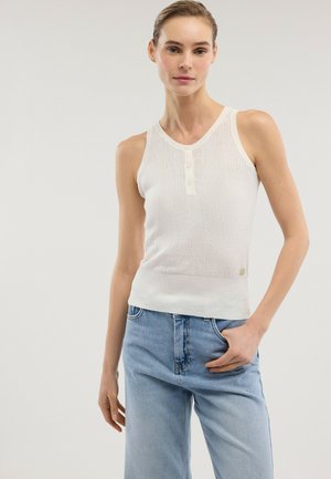 Donna che indossa un top bianco senza maniche a costine con bottoni e jeans azzurri, in piedi con una mano in tasca davanti a uno sfondo uniforme.