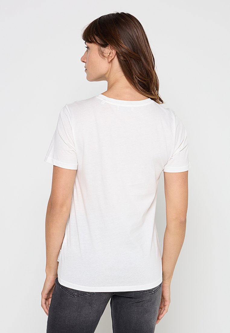 Frau mit braunen Haaren, die ein einfaches weißes T-Shirt mit kurzen Ärmeln und dunkle graue Jeans trägt, mit dem Rücken zur Kamera vor einem schlichten Hintergrund.