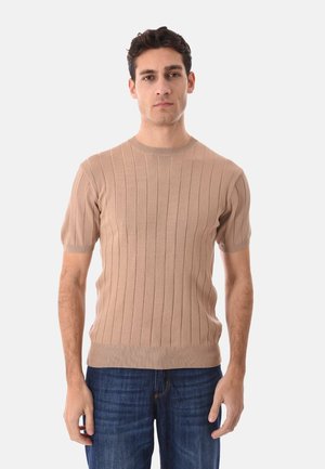 Uomo che indossa un maglione a maniche corte beige a coste e jeans blu, in piedi davanti a uno sfondo semplice grigio chiaro.