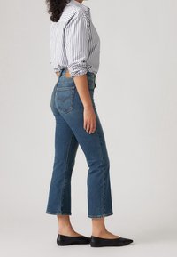 Femme en jean bleu court, chaussures plates noires et chemise blanche à manches longues avec des rayures verticales noires, debout de profil sur un fond uni.