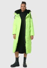 Groene pufferjas met zwarte nepbont kraag, zijzakken, rits aan de voorkant en drukknoopjes. Gedragen over een zwart geribbeld jurk en zwarte laarzen.