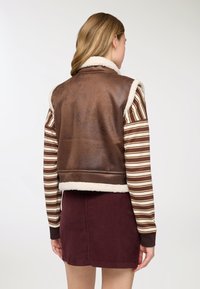 Giacca marrone in pelle ecologica con colletto in shearling, abbinata a una camicia a righe a maniche lunghe e una gonna in velluto a coste color bordeaux. Dettagli di tessuto strutturato visibili.