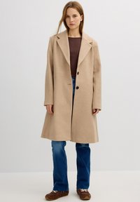 Abrigo de lana beige con solapa muesca, dos botones y bolsillos laterales, combinado con una camiseta marrón de canalé y vaqueros acampanados azules. Zapatos marrones.