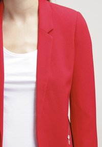 Blazer rouge ajusté en tissu texturé avec une fermeture à un seul bouton et une poche latérale zippée, associé à un top blanc à col rond.