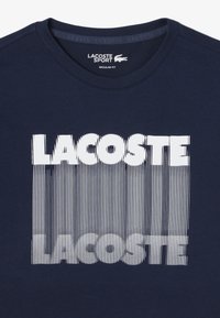 Camiseta de algodón azul marino con un atrevido gráfico blanco que dice "LACOSTE", con patrones de líneas verticales que enfatizan el texto. Diseño de ajuste regular.