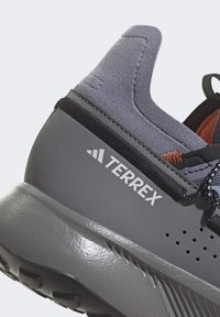 Sapato de caminhada cinza com um upper de camurça texturizado, detalhes em laranja contrastantes e uma sola de borracha moldada com ranhuras de tração.