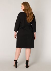 Zwart knie-lengte jurkje met driekwart mouwen, gemaakt van een gladde stof. Het heeft een rechte snit en een detail met een naad in het midden van de rug.