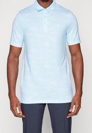 Homme portant un polo bleu clair à manches courtes avec des motifs subtils et un pantalon bleu marine, debout devant un fond uni.
