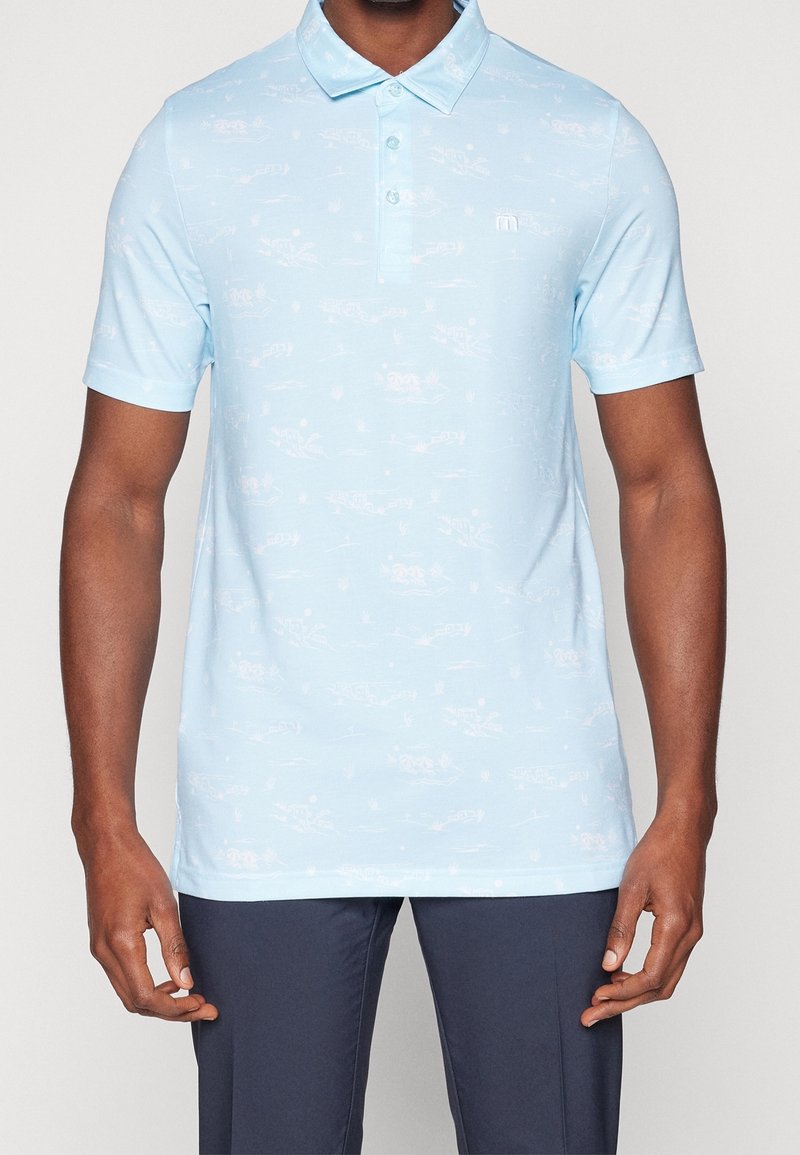 Homme portant un polo bleu clair à manches courtes avec des motifs subtils et un pantalon bleu marine, debout devant un fond uni.