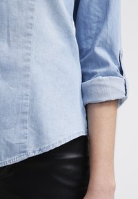 Light blue denim shirt with rolled-up sleeves, featuring a soft texture and a hem that curves slightly. Worn with black pants.