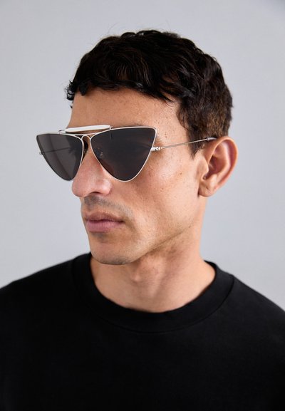 Ochelari de soare cu un cadru metalic argintiu și lentile întunecate, având un design unghiular și o bară proeminentă de sprânceană. Fundal de cămașă neagră.