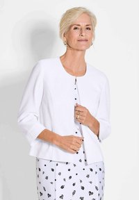 Weißer gerippter Cardigan mit Dreiviertelärmeln, runder Ausschnitt und einzelner Knopfverschluss, getragen über einem schwarz-weißen gemusterten Kleid.