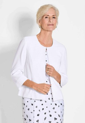 Cardigan blanc côtelé avec des manches trois-quarts, présentant un col rond et une fermeture à bouton unique, porté par-dessus une robe à motifs noir et blanc.