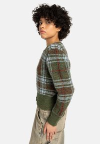 Zöld kockás pulóver kék és piros részletekkel, kötött textúrával, bordás mandzsettákkal és aljjal, rövidített fazonnal, széles nyakkivágással és oldalsó hasítékokkal.