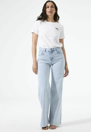 Femme debout portant un t-shirt blanc avec un petit motif de panthère noire et un jean taille haute à jambes larges bleu clair, ainsi que des sandales.