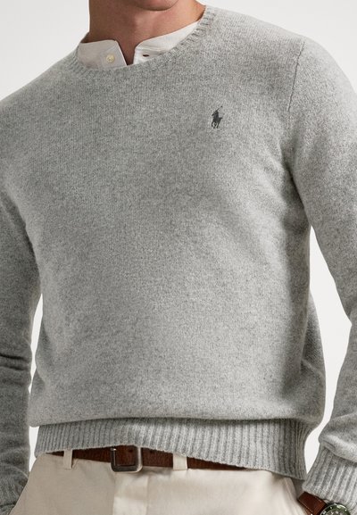 Γκρι πλεκτό πουλόβερ με στρογγυλή λαιμόκοψη, ριμπ τελείωμα και διακριτικό λογότυπο στο στήθος· λεπτή υφή, απαλό ύφασμα. Φορεμένο πάνω από λευκό πουκάμισο με γιακά.