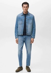 Jachetă din denim albastru deschis cu două buzunare frontale, asociată cu blugi slim-fit asortați. Purtați peste un pulover închis la culoare și pantofi maronii.