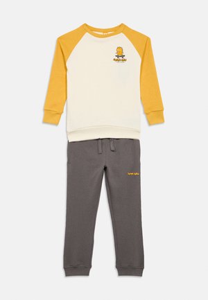 Felpa raglan color crema e senape a maniche lunghe con grafica di dinosaurio su skateboard, abbinata a pantaloni jogger grigi con testo giallo "Skate Crew".