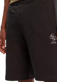 Pantaloni corti neri in misto cotone con una texture morbida, dotati di tasche laterali e ricamo del logo 'GUESS' sul lato sinistro in basso.