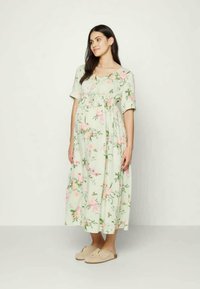 Vestido de maternidade floral em verde claro com mangas curtas, corpete franzido e comprimento até os tornozelos. Apresenta padrões florais em rosa e branco.