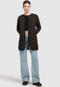 Zwarte gevoerde lange jas met een ronde halslijn, rits aan de voorkant en een getextureerd ruitpatroon. Gecombineerd met lichtblauwe wijde jeans en zwarte sneakers.