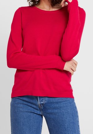 Rotes langärmeliges Oberteil aus Rippstoff, mit Rundhalsausschnitt und enganliegenden Bündchen. Getragen mit blauen Jeans; enthält ein kleines Logodetail.