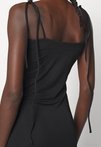 Vue de dos d'une personne aux cheveux tressés portant une robe noire sans manches avec de fines bretelles nouées aux épaules.