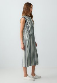 Gestreiftes, ärmelloses Kleid mit blauen und weißen vertikalen Mustern. Fließende Silhouette, knielang, kombiniert mit weißen Sneakers. Weicher Stoff.