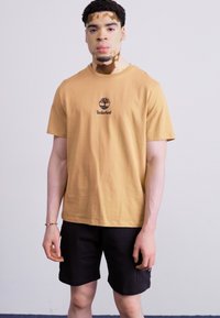 Brun bomulls t-skjorte med svart Timberland-logo, kortermet, kombinert med svarte shorts. Modellen står foran en enkel lys bakgrunn.