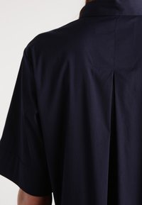 Mörkblå skjorta med krage, korta ärmar och en vecket detalj bak, tillverkad av slätt tyg med en lätt lyster.