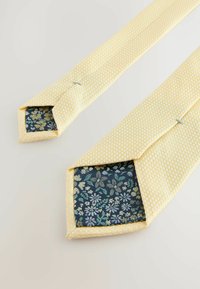 Gul slips med små polka prikker. Indersiden er mørkeblå med et blomstermønster i grøn og lyseblå. Glat tekstur, spidse ender.