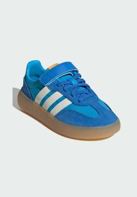 Blue athletic shoe with suede and synthetic materials, featuring white stripes, lace-up design, Velcro strap, and a rubber sole.