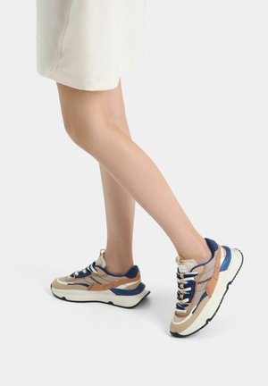 Jambes dans une jupe beige portant des baskets chunky beige, bleues et blanches, montrées en mi-pas contre un fond gris clair uni.