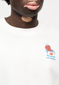 Λευκό βαμβακερό φούτερ με πολύχρωμο γραφικό που απεικονίζει έναν χαμ ογελαστό αστακό να βγαίνει από ένα κουτί με την επιγραφή "new balance".